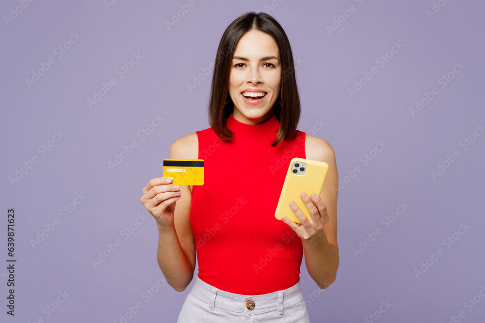 Young woman wears red tank shirt casual clothes using mobile cell phone hold credit bank card doing online shopping order delivery booking tour isolated on plain pastel light purple background studio. Young woman wears red tank shirt casual clothes using mobile cell phone hold credit bank card doing online shopping order delivery booking tour isolated on plain pastel light purple background studio.