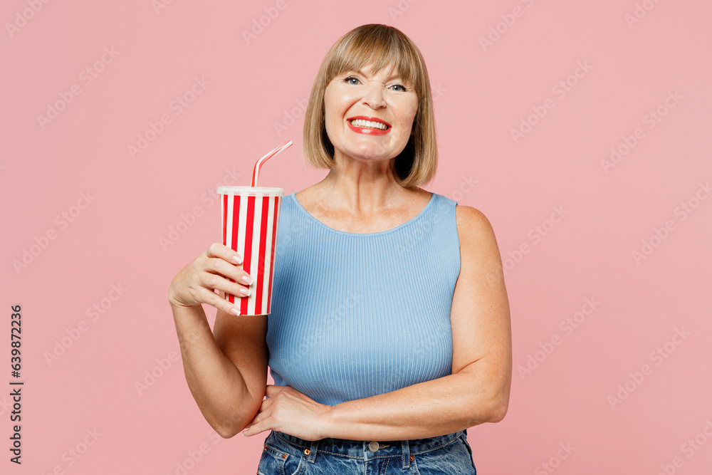 Elderly fun blonde woman 50s years old she wears blue undershirt casual clothes hold in hand cup of soda pop cola fizzy water isolated on plain pastel light pink background studio. Lifestyle concept. Elderly fun blonde woman 50s years old she wears blue undershirt casual clothes hold in hand cup of soda pop cola fizzy water isolated on plain pastel light pink background studio. Lifestyle concept.