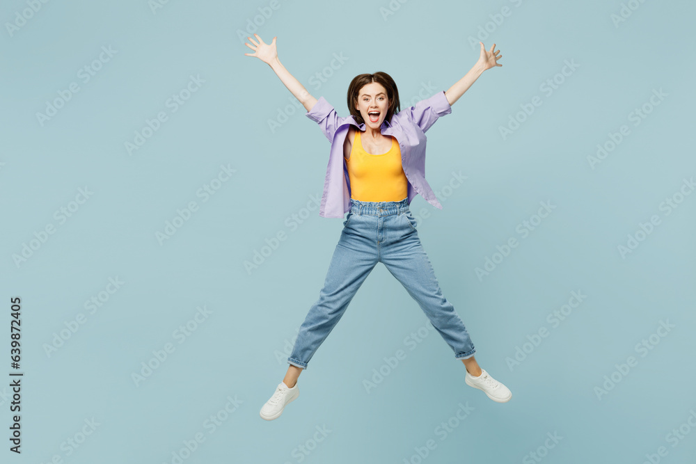 Full body young happy woman she wears purple shirt yellow t-shirt casual clothes jump high with outstretched hands look camera isolated on plain pastel light blue background studio. Lifestyle concept.