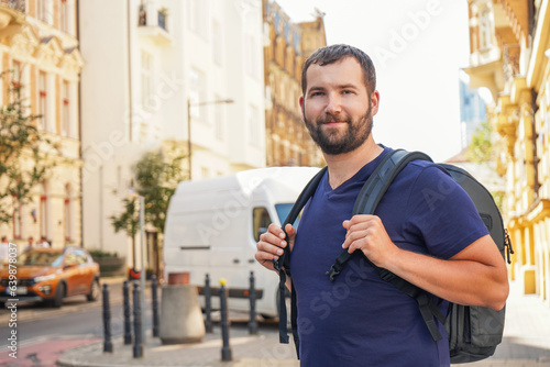 a handsome bearded man with a backpack in the european center looks at the camera and smiles against the backdrop of beautiful buildings, architecture.Tourism, summer holidays, backpack