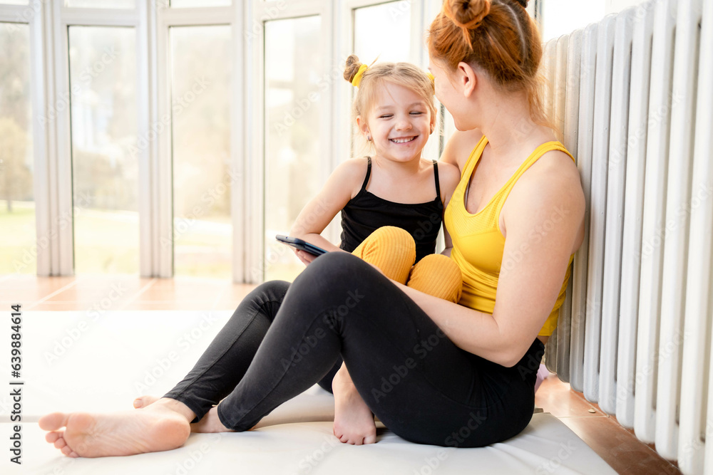 Young fit mom having loving time with her daughter, looking at a phone together