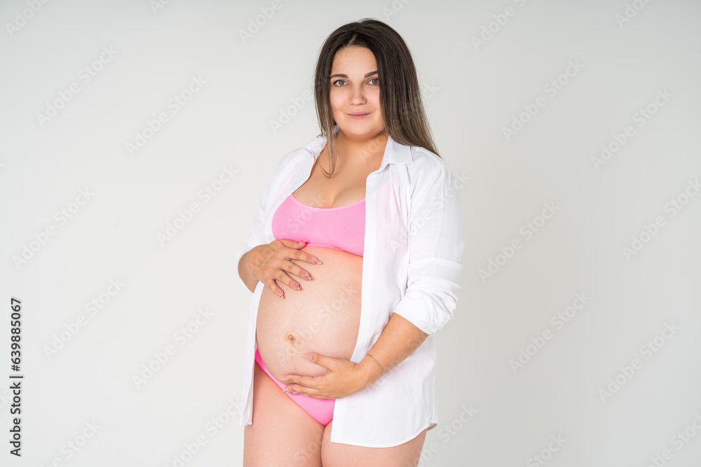 Happy plus size pregnant woman in pink underwear on gray background, maternity clothes, pregnancy concept Happy plus size pregnant woman in pink underwear on gray background, maternity clothes, pregnancy concept