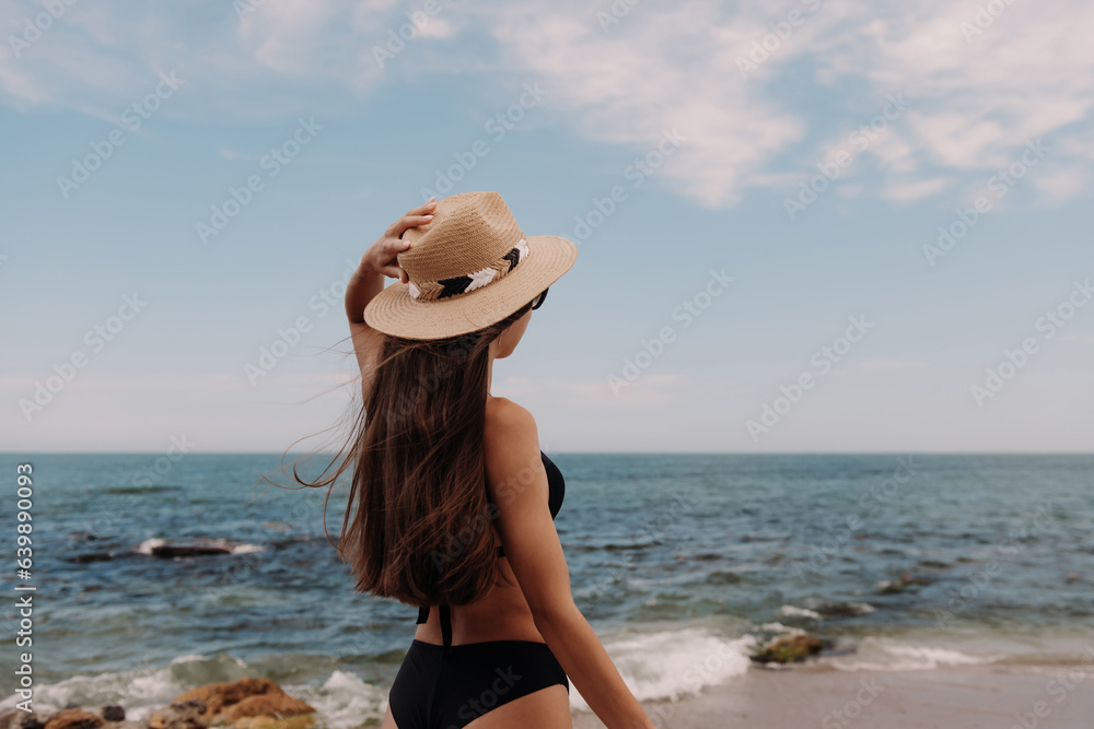 Rear view of beautiful woman in swimwear adjusting hat while enjoying sea view on the beach