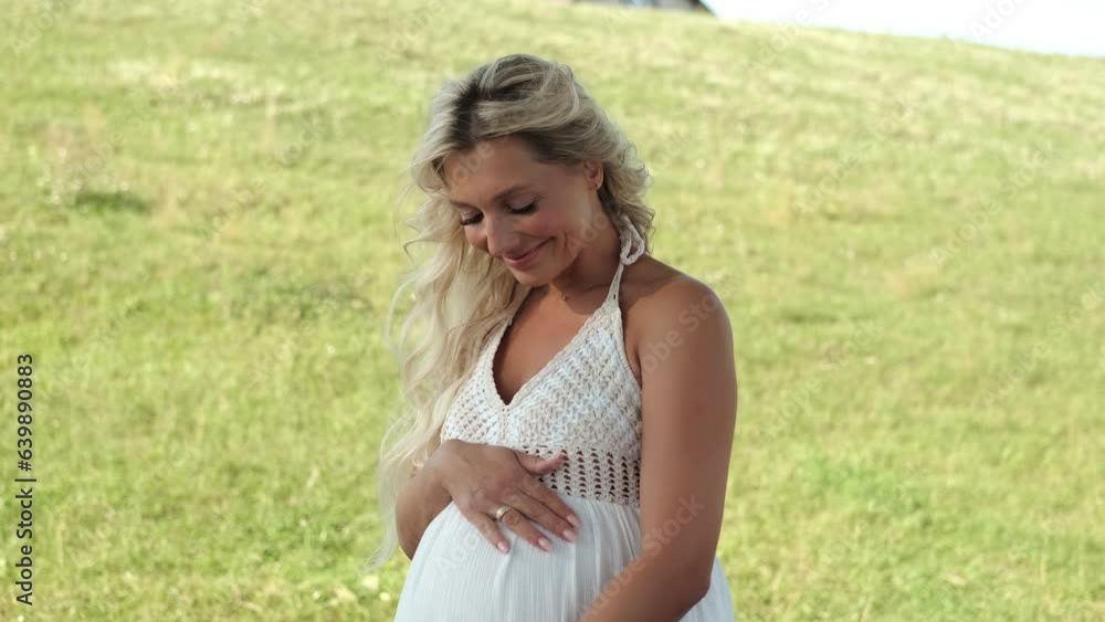 A pregnant woman in a white dress strokes her belly and smiles while standing on the lawn. A pregnant woman in a white dress strokes her belly and smiles while standing on the lawn.