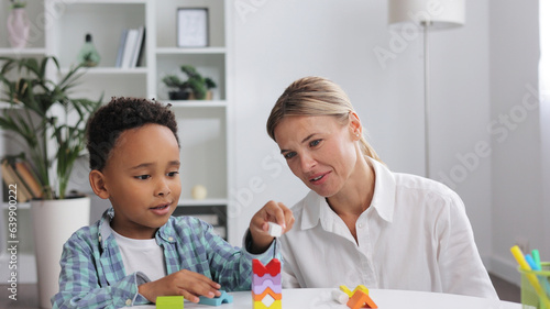 African American child curly little boy and child development specialist blonde woman playing with colorful wooden bricks sitting at table, making pyramid together. Exercises for children with autism.