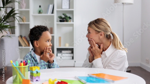 Cute little multiracial boy with speech problems practicing with occupational therapist, practicing and looking at mirror in office. The concept of childhood autism.