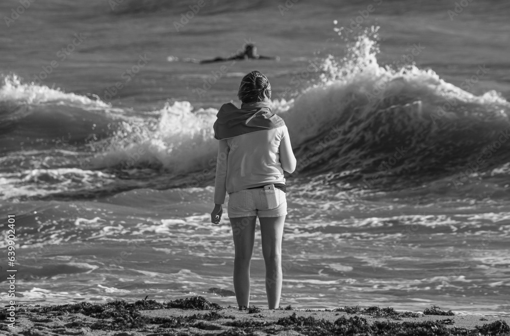 Fototapeta premium A woman on a Vendée beach watches surfers on the ocean, dressed in shorts with a smartphone in the back pocket, a small towel on her back, black and white photo.