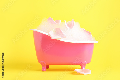 Small bathtub with foam and rose petals on yellow background