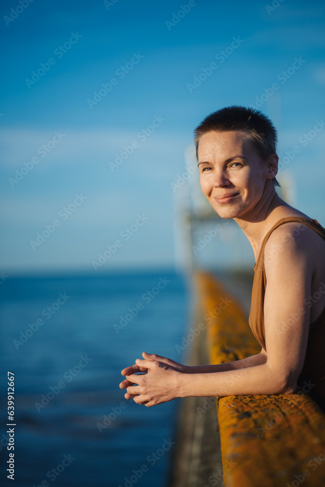 A middle-aged woman stands on a pier in the sunset rays on the ocean or sea. Peaceful inner joy. Weekends, holidays, rest. Happy face.