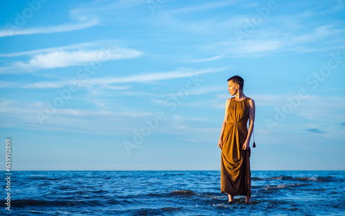 A woman on the ocean looks away. Woman with short hair and long dress. Blue ocean and blue sky.