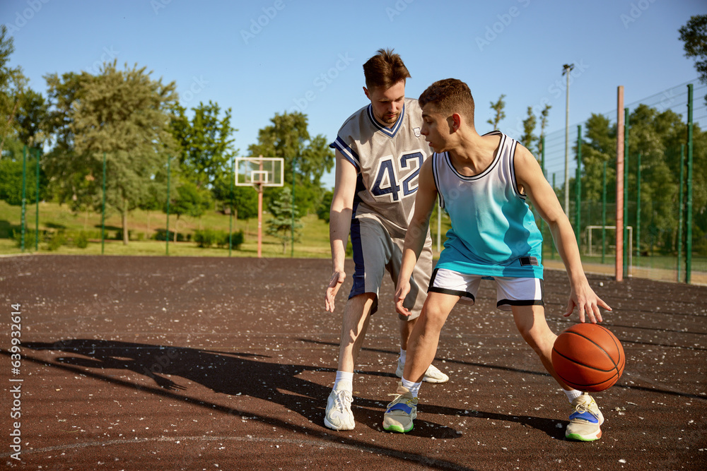 Young trainer teaching teenage boy playing basketball on street court
