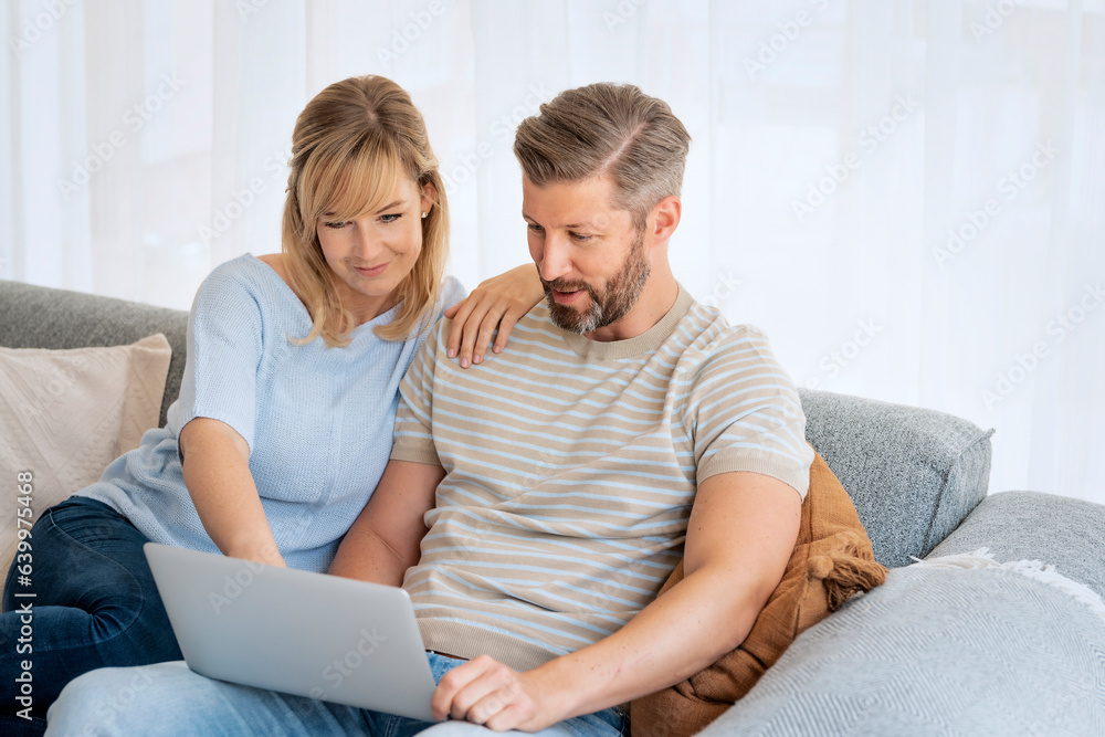 Middle-aged couple wearing casual clothes and sitting at home on the sofa and using a laptop Middle-aged couple wearing casual clothes and sitting at home on the sofa and using a laptop
