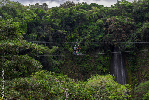 couple of hikers with harness and helmet riding zipline bicycles surrounded by vegetation and a beautiful waterfall in Costa Rica