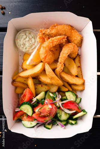 Shrimp fried breaded with lemon, rustic potato wedges, vegetable salad and sauces in a paper box.