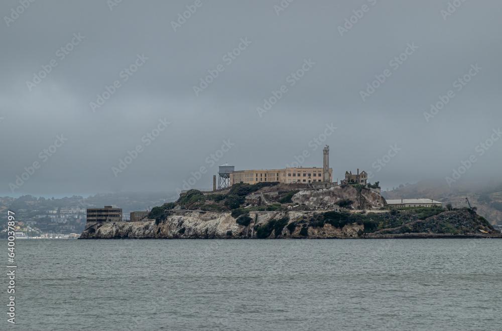 San Francisco, CA, USA - July 12, 2023: Alcatraz Island with Federal Prison monumental museum under foggy morning sky. Drak green vegetation on gray rocks. Gray Bay water.