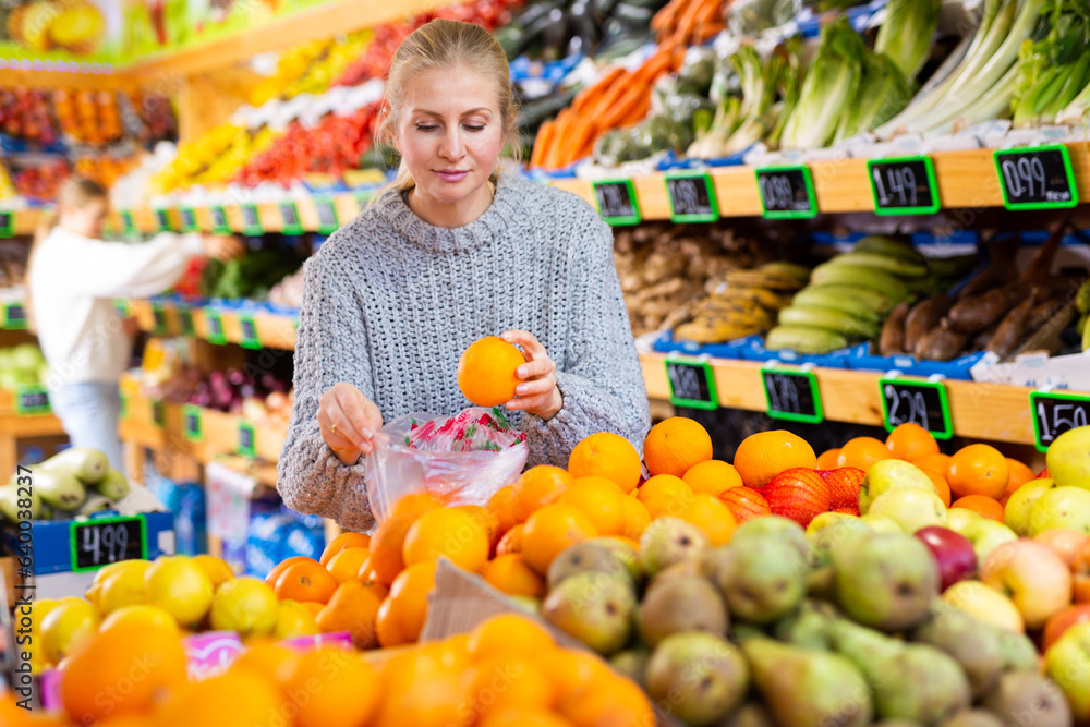 Smiling woman shopping for organic fruits in farmer store, choosing sweet ripe oranges ..