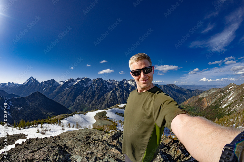 Adventurous athletic male hiker taking a selfie with the sun shinning and rugged mountains in the background in the Pacific Northwest.
