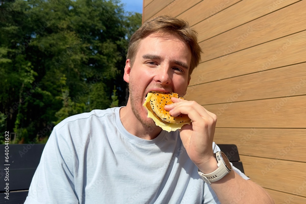 Portrait of young happy man is eating, biting juicy delicious burger outside cafe looking at camera and smile. Fast junk food, unhealthy eating concept. Portrait of young happy man is eating, biting juicy delicious burger outside cafe looking at camera and smile. Fast junk food, unhealthy eating concept.