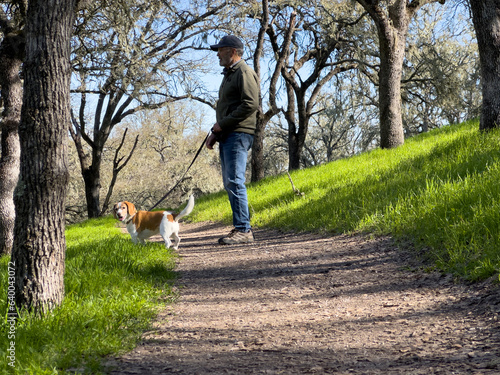 Man with ball cap and jeans standing in middle of dirt path with small, cute beagle on leash, looking at the camera