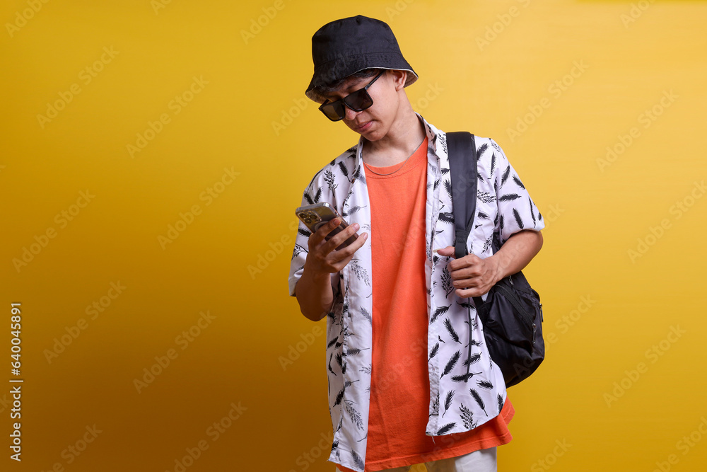 Young Asian man looking at mobile phone while standing against yellow background carrying bacpkack Young Asian man looking at mobile phone while standing against yellow background carrying bacpkack