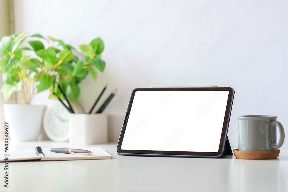 Digital tablet with blank display, coffee cup and potted plant on white table. Simple workplace.