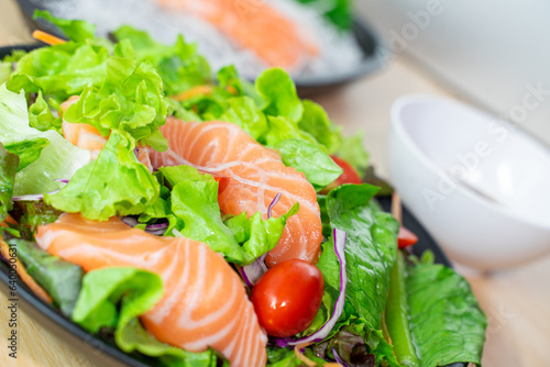 Salmon salad contains to raw salmon with lettuce, tomato and red cabbage. which puts on nature-wooden table. Blur another objects on background.