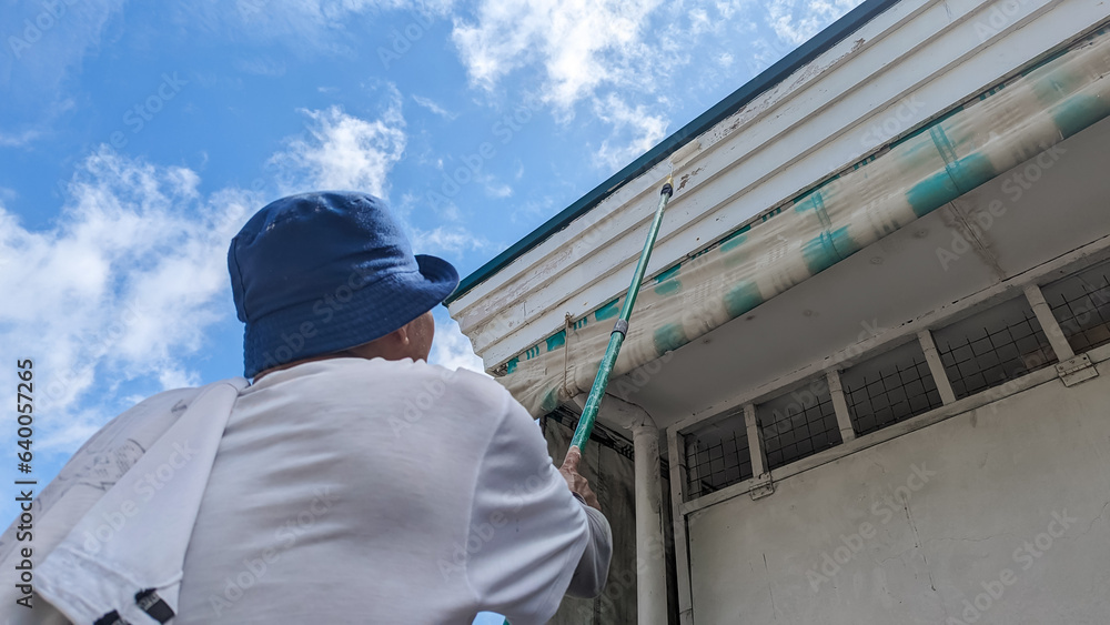 A handyman paints the eaves of a small store using a paint roller attached to a long pole. Small business shop exterior renovation.