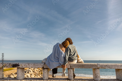 Rear view of couple hugs while sitting on bench and enjoys beautiful ocean view. High quality photo