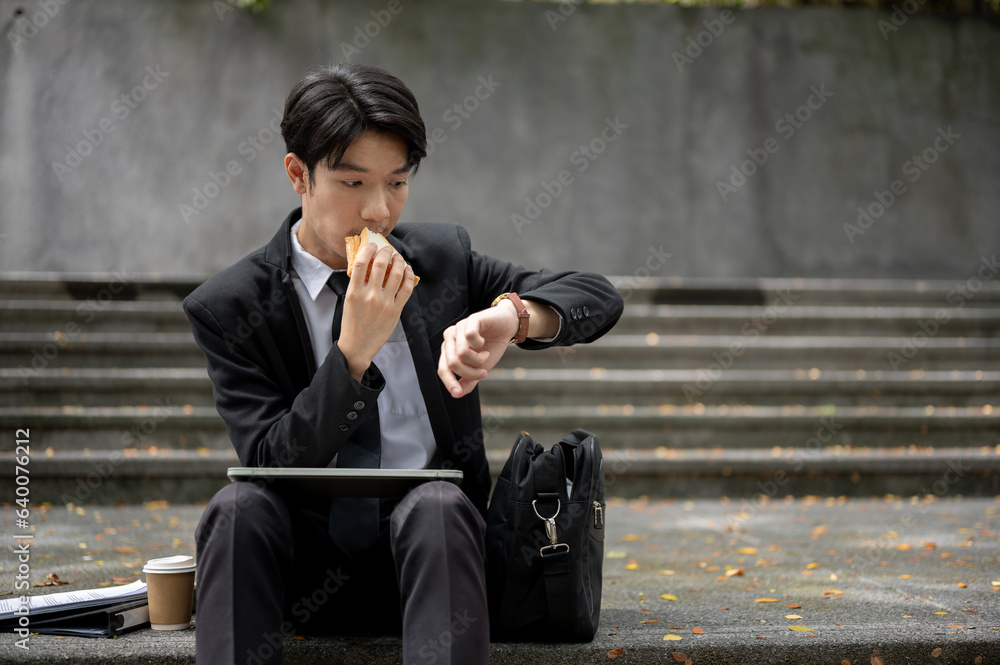 A busy and hungry Asian businessman is checking time on his watch and eating a sandwich