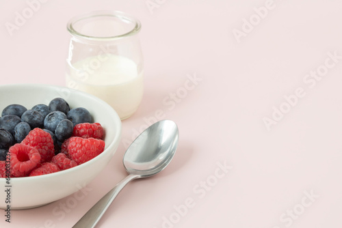 Closeup view of a white bowl with berries, glass of milk and a spoon on light pink background with copy space