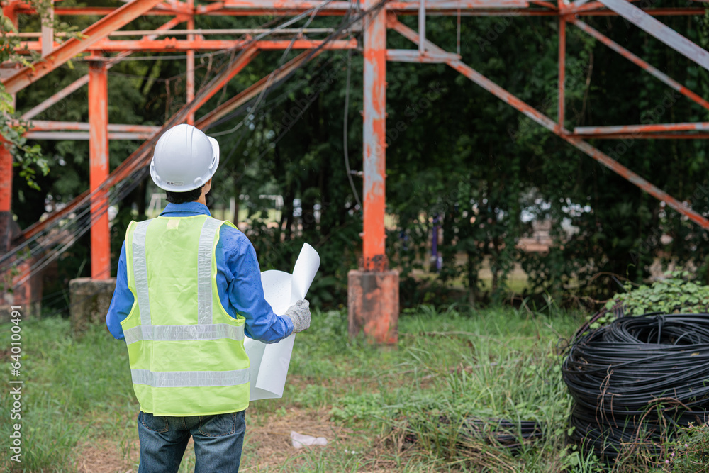 The electrical engineer wears a blue shirt overlaid with green, wears white pieces, and wears protective goggles. Checking operation, holding model in hand