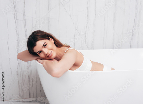 Portrait of young beautiful sexy woman in white lingerie. Smiling carefree model wearing pure underwear. Hot brunette posing in bath interior. Perfect body. Female in luxury bathroom