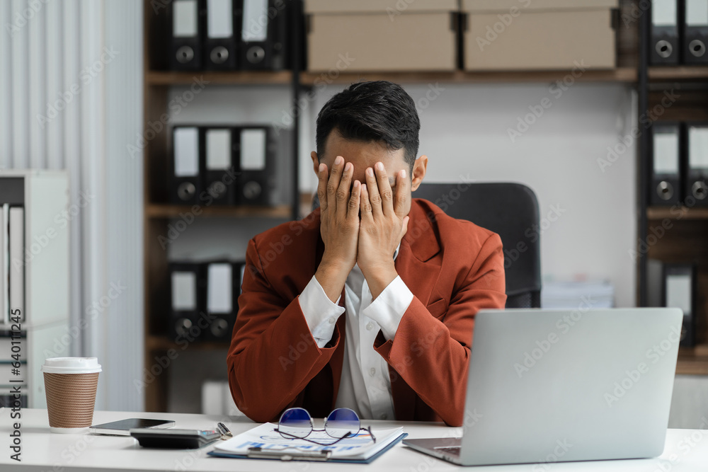 Young sad businessman is sitting at table, covering his face with his hand. On desk is laptop, tablet computer, Stress. Young sad businessman is sitting at table, covering his face with his hand. On desk is laptop, tablet computer, Stress.