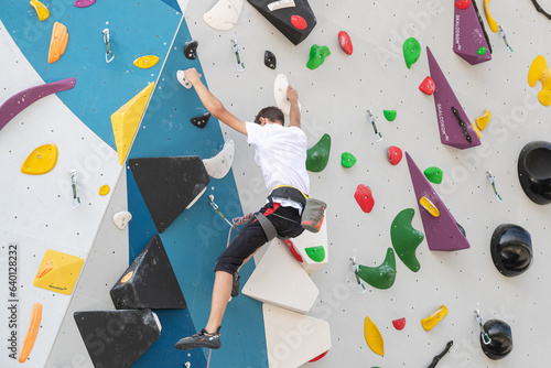 A climber climbs an artificial stone wall using a harness. Active young guy on a stone wall in a sports center. The guy trains on a rock climber outdoors. A young, athletic man climbs a climbing wall.