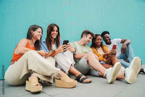 Fotografija Multiracial smiling teenagers having fun sharing on the social media with their smart phones sitting in row on the floor, blue background