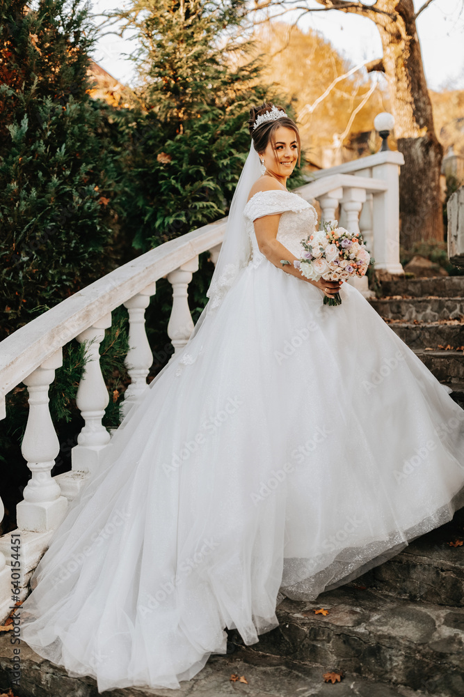 Fototapeta premium Portrait of the bride in the autumn park on stone steps. The bride in a wedding dress on a natural background with a bouquet of flowers in her hands. Wedding day.