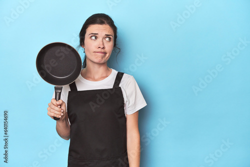 Woman with apron and pan on blue background confused, feels doubtful and unsure.