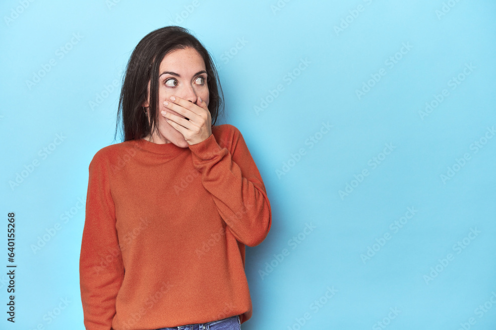 Young caucasian woman on blue backdrop thoughtful looking to a copy space covering mouth with hand. Young caucasian woman on blue backdrop thoughtful looking to a copy space covering mouth with hand.