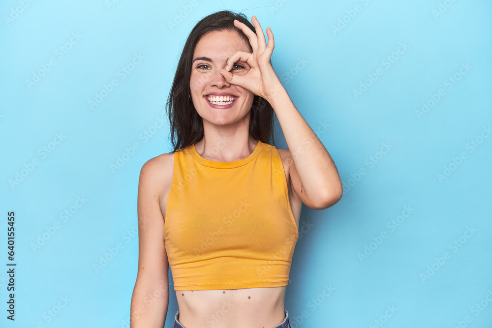 Young caucasian woman on blue backdrop excited keeping ok gesture on eye. Young caucasian woman on blue backdrop excited keeping ok gesture on eye.