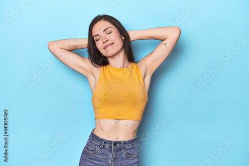 Young caucasian woman on blue backdrop feeling confident, with hands behind the head.