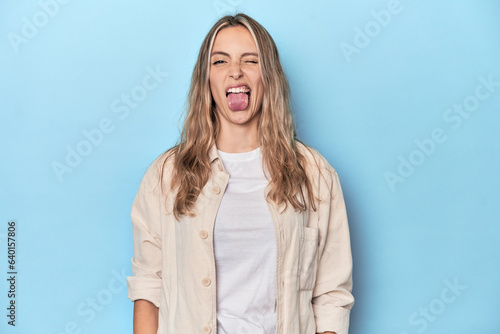 Blonde young caucasian woman in blue studio funny and friendly sticking out tongue.