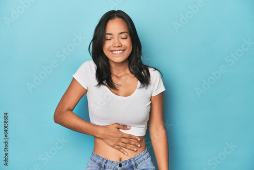 Filipina young woman on blue studio touches tummy, smiles gently, eating and satisfaction concept.