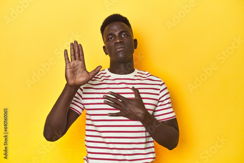 Stylish young African man on vibrant yellow studio background, taking an oath, putting hand on chest.