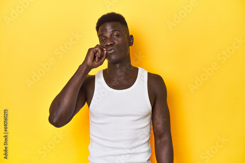 Stylish young African man on vibrant yellow studio background, with fingers on lips keeping a secret.