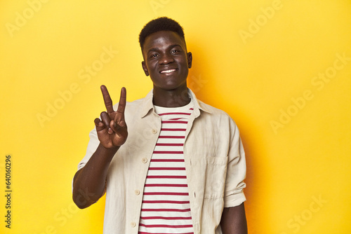 Stylish young African man on vibrant yellow studio background, showing victory sign and smiling broadly.