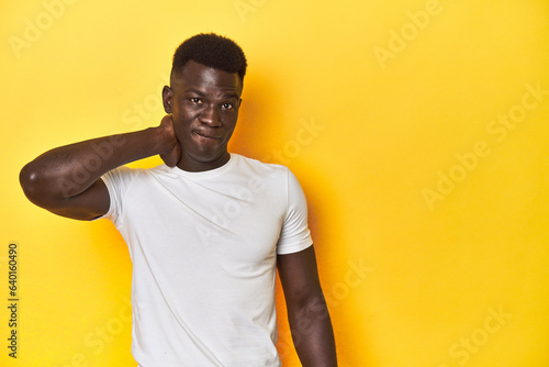 Stylish young African man on vibrant yellow studio background, touching back of head, thinking and making a choice.
