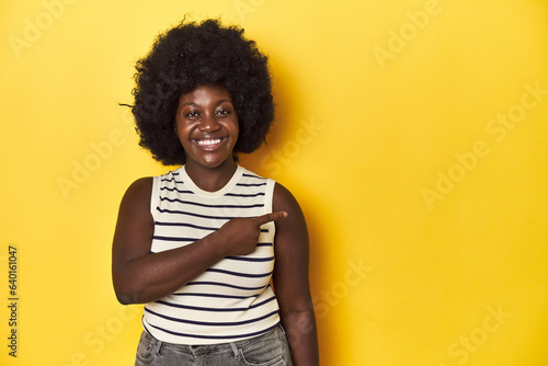 African-American woman with afro, studio yellow background smiling and pointing aside, showing something at blank space. © Asier