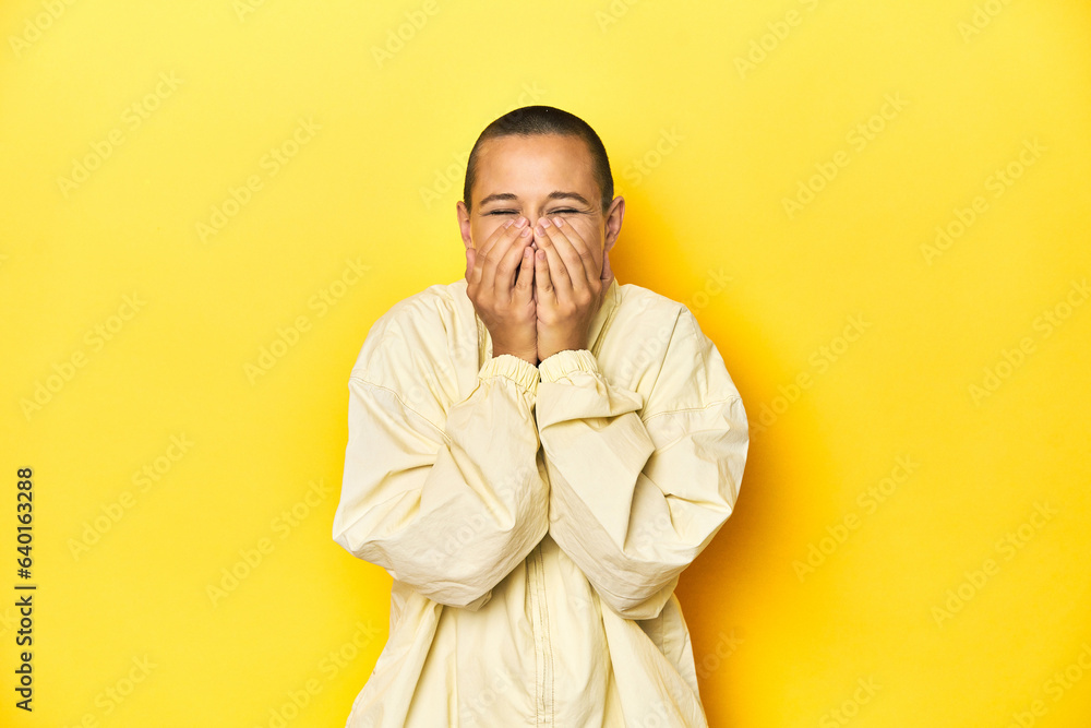 Fototapeta premium Young woman in jacket, yellow studio backdrop laughing about something, covering mouth with hands.