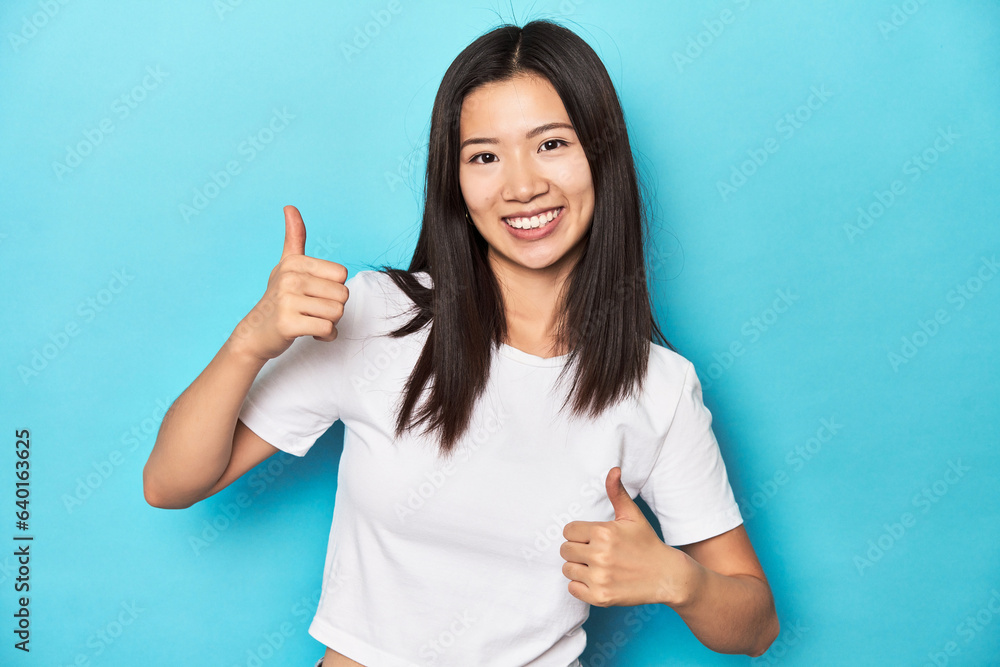 Fototapeta premium Young Asian woman in white t-shirt, studio shot, raising both thumbs up, smiling and confident.