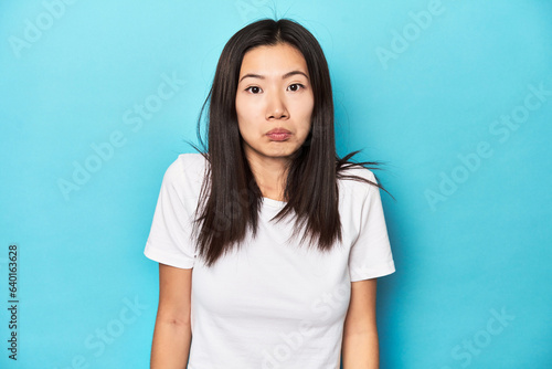 Young Asian woman in white t-shirt, studio shot, shrugs shoulders and open eyes confused.